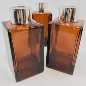 May include: Three amber glass bottles with silver tops. The bottles have a rectangular shape with angled sides. The tops are cylindrical and metallic. The bottles are arranged in a group, with two in the foreground and one in the background.