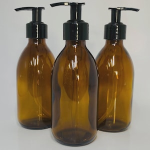 May include: Three amber glass soap dispensers with black pump tops. The bottles have a rounded shape and are filled with a clear liquid. The dispensers are arranged in a row against a white background.