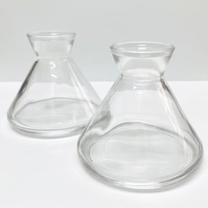 May include: Two clear glass vases with a conical body and a flared, rounded top. The vases are empty and sit on a white surface. The glass is transparent, allowing a clear view of the interior.