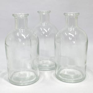 May include: Three clear glass bottles with rounded bodies and narrow necks. The bottles are empty and transparent, allowing a view of the background. The bottles are arranged closely together, suggesting a set or collection. The glass has a slight sheen.