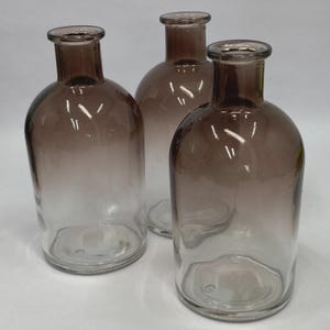 May include: Three clear glass bottles with a brown ombre effect. The bottles have a rounded shape with a narrow neck and a flat base. The glass is transparent at the bottom and fades to brown at the top.