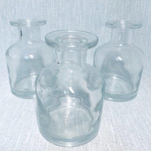 May include: Three clear glass bud vases with a rounded body and a short, wide neck. The vases are empty and transparent, allowing light to pass through. The vases are arranged on a light-coloured surface.