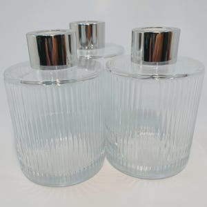 May include: Three clear glass diffuser bottles with silver-coloured caps. The bottles have a ribbed design and a round shape. The caps are shiny and reflect light. The bottles are empty and ready to be filled with fragrance.