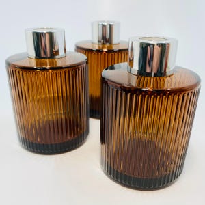 May include: Three amber glass diffuser bottles with vertical ribbed detailing. Each bottle has a silver cap. The bottles are cylindrical with a rounded top and base. The glass has a gradient from light to dark amber.