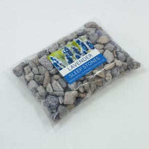 May include: A transparent bag containing small, grey and purple stones. The label reads "LAVENDER SLEEP STONES MADE WITH LAVENDER ESSENTIAL OIL" with a lavender flower image.