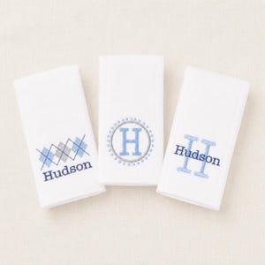 May include: Three white rectangular towels featuring blue and grey embroidery. The left towel has an argyle pattern and the name Hudson. The center towel displays a letter H inside a dotted circle. The right towel shows the name Hudson with two large H letters.