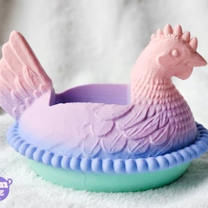 Puede incluir: Un recipiente en forma de gallina en colores pastel. La gallina es rosa, morada y azul, con un dise&ntilde;o detallado de plumas. La base es verde claro con un borde azul. El logotipo de Cluckin' Goodz es visible.