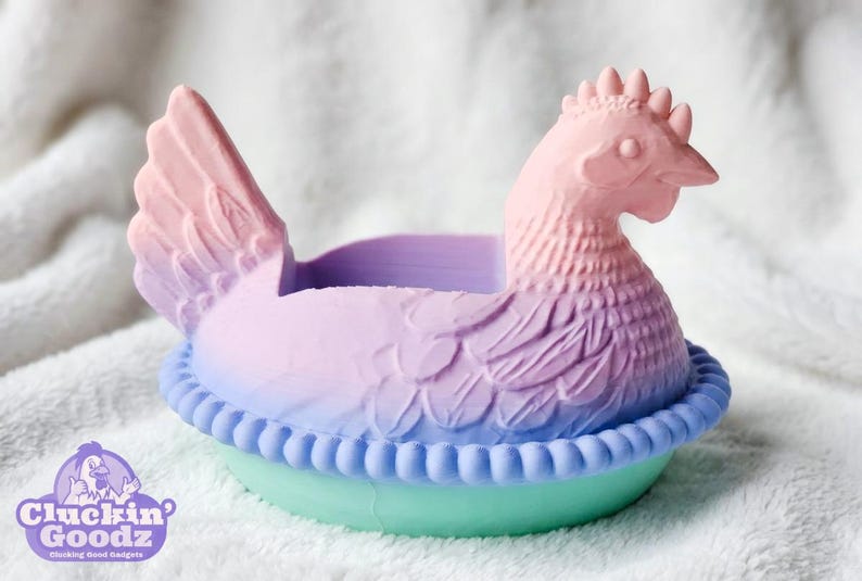 Puede incluir: Un recipiente en forma de gallina en colores pastel. La gallina es rosa, morada y azul, con un dise&ntilde;o detallado de plumas. La base es verde claro con un borde azul. El logotipo de Cluckin' Goodz es visible.