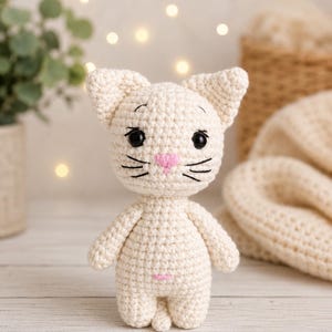 May include: A small, cream-colored crocheted cat figurine with a round head, pointed ears, and a compact body. It features black plastic safety eyes, embroidered black eyebrows and whiskers, a small pink stitched nose, and a tiny pink belly mark.