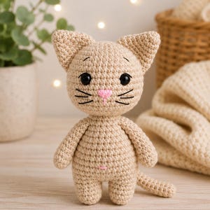 May include: A beige crochet cat doll featuring a round head with pointed ears, large black plastic eyes, a small pink embroidered nose, and stitched whiskers. The body is compact with short arms, two legs, and a small tail, all in a matching beige yarn.