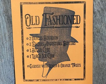 May include: A vintage-style orange poster with black text and a silhouette of a top hat. The text reads "OLD FASHIONED" and lists the ingredients for an Old Fashioned cocktail: 2 ounces bourbon, 2 dashes Angostura bitters, 1 sugar cube, 1 large ice cube, and garnish with lemon and orange twists.