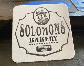 May include: White square coaster with a black and white logo for Solomons Bakery in Camden Town, London. The logo features a stylized image of two people wearing hats.