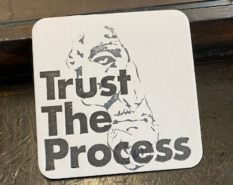 May include: A white coaster with a black and white image of a person's face and the text "Trust The Process".