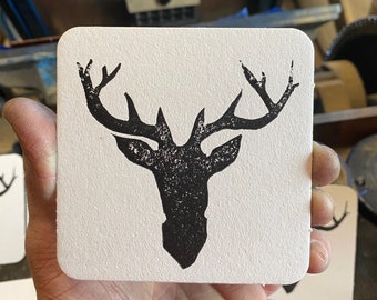May include: A white square coaster with a black silhouette of a deer head with antlers.