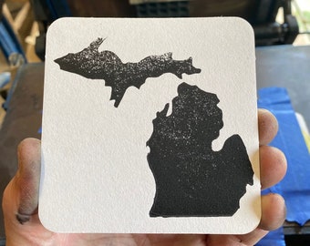 May include: A black and white silhouette of the state of Michigan printed on a white coaster.