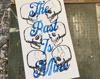 May include: A white print with blue text that reads "The Past Is Mine". The text is surrounded by five black and white skull illustrations.
