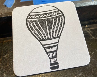 May include: A black and white illustration of a hot air balloon on a white coaster.