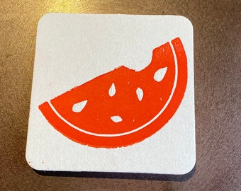 May include: A white square coaster with a red watermelon slice design. The watermelon slice has black seeds.