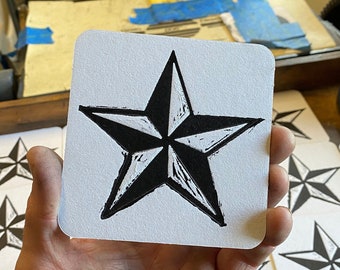 May include: A hand holds up a white coaster with a black and white nautical star design. The coaster is one of many on a wooden table.
