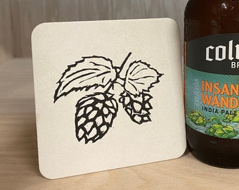 May include: A brown glass bottle of Columbus Brewing Co. Insane Wanderer India Pale Ale. The bottle has a white label with black and orange text. The label features a cartoon illustration of a hop plant.