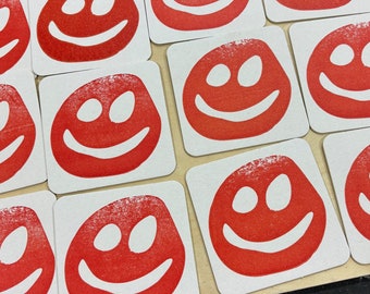 May include: A set of 12 white square coasters with red smiley faces printed on them. The smiley faces have two eyes and a wide smile.