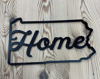 May include: A black metal sign in the shape of Pennsylvania with the word "Home" in cursive script. The sign is mounted on a light-colored wooden surface with visible wood grain.