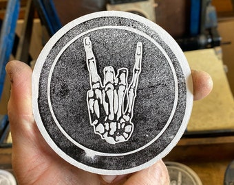 May include: A black and white round coaster with a skeleton hand making the rock and roll hand gesture. The coaster is being held by a hand.