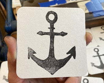 May include: A close-up of a white coaster with a black anchor design. The anchor is a simple design with a rounded top and two points at the bottom. The coaster is part of a set of coasters with the same design.