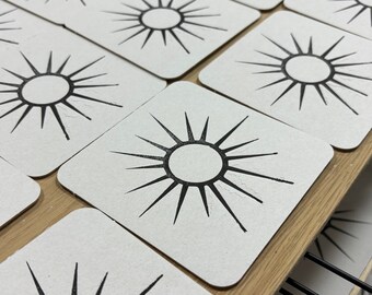 May include: A collection of square coasters with a black sunburst design on a white background. The coasters are arranged on a wooden shelf, showcasing a minimalist and geometric aesthetic. The sunburst design features a central circle with radiating lines.