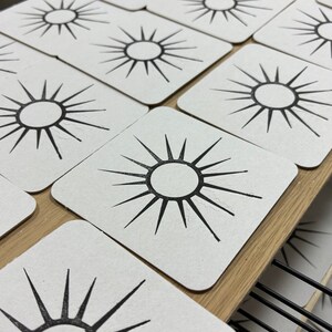 May include: A collection of square coasters with a black sunburst design on a white background. The coasters are arranged on a wooden shelf, showcasing a minimalist and geometric aesthetic. The sunburst design features a central circle with radiating lines.