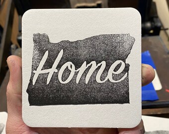 May include: A white coaster with a black outline of the state of Oregon and the word "Home" printed in white inside the outline.