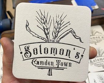 May include: Square white coaster with a black wheat stalk design and the text "Solomon's Camden Town". The coaster is held in a hand, with other coasters visible in the background. The image is taken in a workshop.