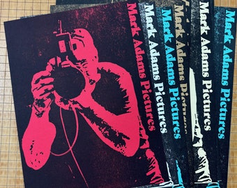 May include: A stack of five black and white posters with the text "Mark Adams Pictures" printed in various colors, including red, blue, and yellow. The posters feature a silhouette of a person holding a camera.