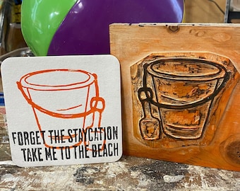 May include: A wooden block with a carved image of a bucket and shovel. A white coaster with a black and orange illustration of a bucket and the text "Forget the staycation take me to the beach" is in the foreground. A purple and green beach ball is in the background.