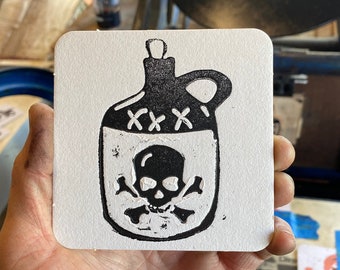 May include: A white square coaster with a black and white linocut print of a jug with a skull and crossbones. The jug has a handle and a stopper. The skull and crossbones are inside the jug.