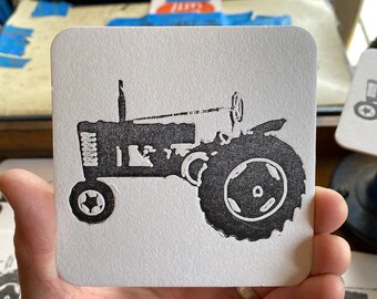 May include: A black and white image of a vintage tractor on a white coaster. The tractor has a large front tire and a smaller back tire.