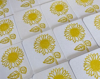 May include: A set of 14 white coasters with a yellow sunflower design. The sunflowers are outlined in black and have a yellow center.