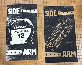 May include: Two black and white printed cards with the word "SIDE" and three stars above the word "ARM". The left card features a Stanley PowerLock 12' tape measure. The right card features a Leatherman multi-tool.
