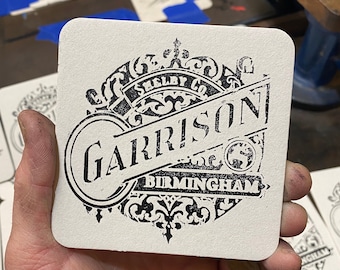 May include: Square, off-white coaster with a black, vintage-style design. The design includes the word "Garrison" in a bold font, with "Birmingham" below. The coaster is held in a hand, with other coasters visible.