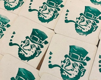 May include: A set of 10 square coasters featuring a green silhouette of a leprechaun with a pipe and a hat. The coasters are made of a light brown material.