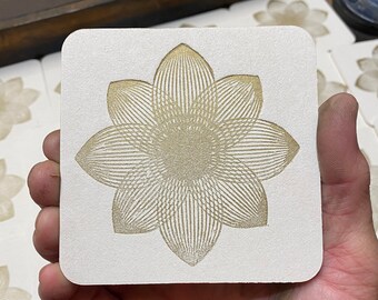 May include: A white square coaster with a gold geometric flower design.