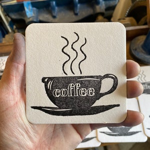 May include: A white coaster with a black and white illustration of a coffee cup with steam rising from it. The text "coffee" is printed on the cup.