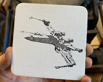 May include: A white coaster with a black and white image of a spaceship. The spaceship is in a flying position with its wings spread out.