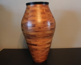 May include: A large, wooden vase with a black rim. The vase is made of many small pieces of wood that are arranged in a pattern. The vase is a light brown color.