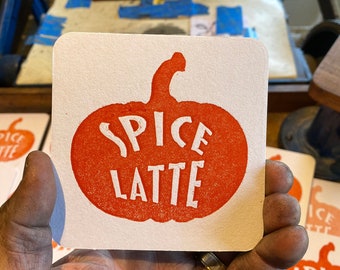 May include: A white coaster with an orange pumpkin design and the text "SPICE LATTE" in white.