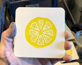 May include: A square, white coaster with a yellow lemon slice design. The lemon slice is hand-printed with a white interior and a yellow rind. The coaster is held in a hand, with other coasters visible in the background.