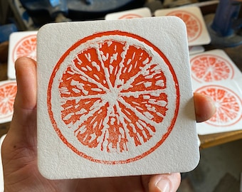 May include: A white square coaster with a red orange slice design printed on it. The coaster is made of a textured paper material.