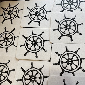 May include: A set of ten square coasters with a black and white printed design of a ship's wheel.
