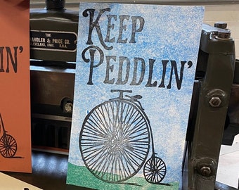 May include: Three vintage-style prints with the text "Keep Peddlin'" and a black and white illustration of a bicycle with a large front wheel. The prints are on different colored paper: orange, blue, and cream.