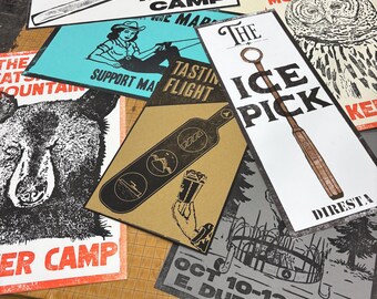 May include: A collection of screen-printed posters with various designs and text. One poster features a hammer and the words "FORGING AHEAD AT MAKER CAMP." Other posters include an owl, a bear, and the text "THE CATSKILL MOUNTAIN." Another poster says "THE ICE PICK."